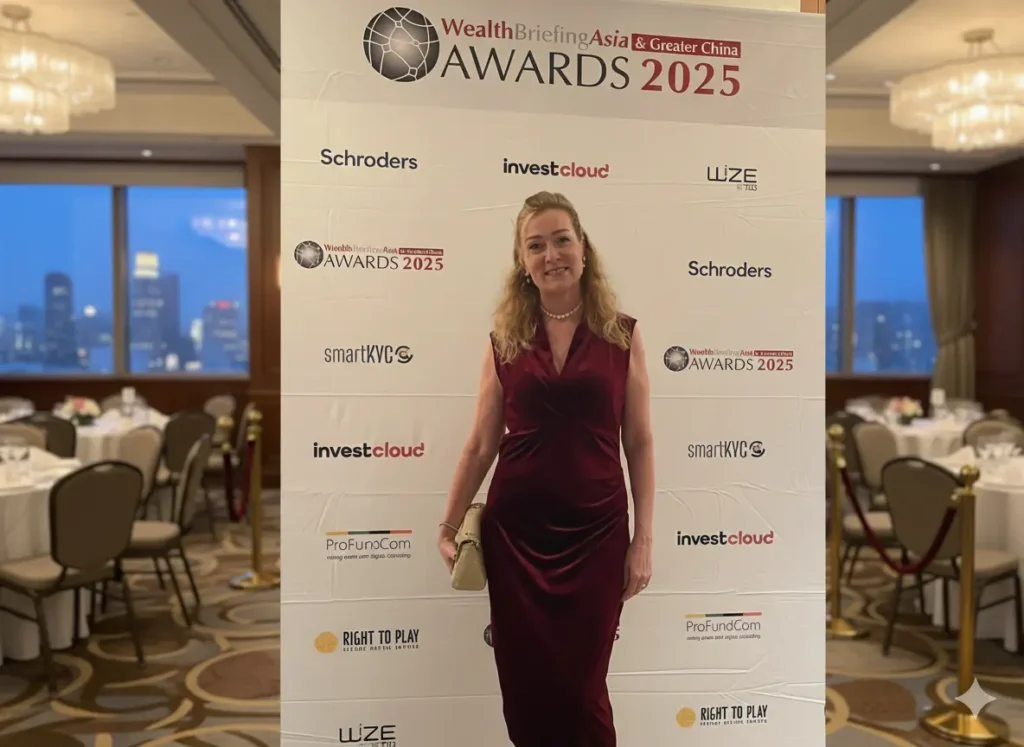 Wealth Briefing Asia Awards 2025, Woman in a burgundy velvet evening gown standing in front of a WealthBriefingAsia Awards 2025 step-and-repeat banner at a formal event venue.