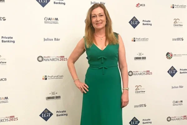 A woman in a green dress and gold shoes poses with her hand on her hip, standing in front of a large event banner for the "WealthBriefingAsia EAM AWARDS 2025" sponsored by LGT Private Banking, Julius Bär, and others.