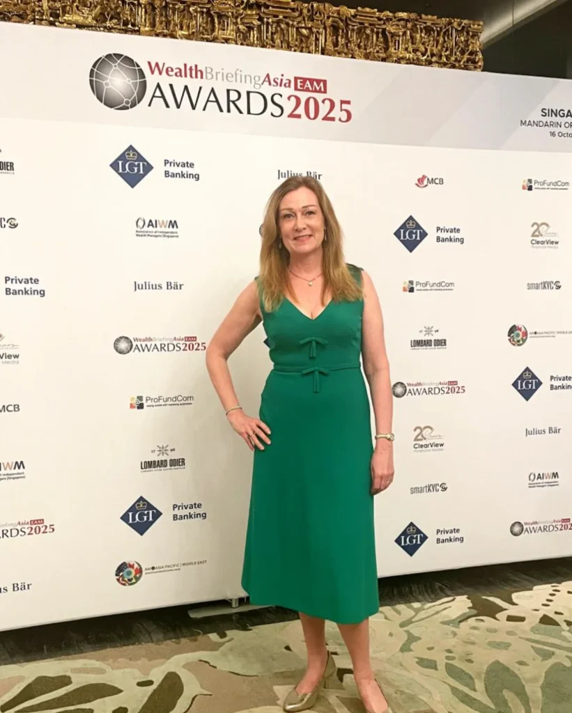 A woman in a green dress and gold shoes poses with her hand on her hip, standing in front of a large event banner for the "WealthBriefingAsia EAM AWARDS 2025" sponsored by LGT Private Banking, Julius Bär, and others.
